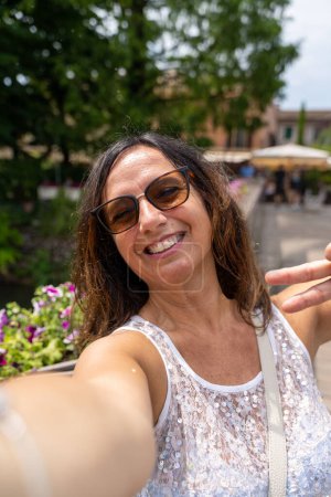 beautiful middle aged woman takes a selfie outdoors on a lakeside terrace - travel and vacation concept