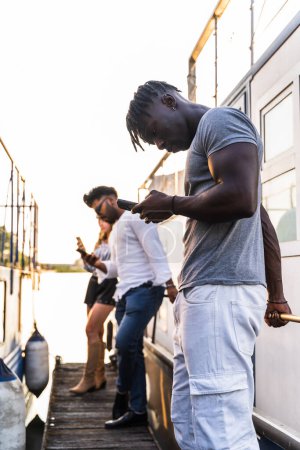 Foto de Pequeño grupo de amigos multiétnicos que se divierten con el teléfono inteligente - concepto de vacaciones de verano en un barco que navega en un río - Imagen libre de derechos