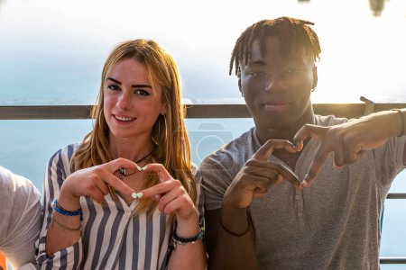 Téléchargez les photos : Petit groupe de jeunes amis multiethniques adultes montrant signe du cœur avec les mains concept de vacances d'été sur un navire sur la rivière - en image libre de droit