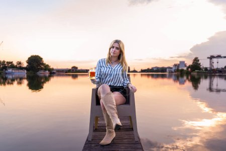 Téléchargez les photos : Jeune femme blonde buvant de la bière assise sur le quai d'un port fluvial au coucher du soleil - en image libre de droit