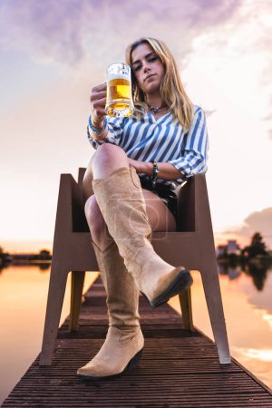 Téléchargez les photos : Jeune femme blonde buvant de la bière assise sur le quai d'un port fluvial au coucher du soleil - en image libre de droit