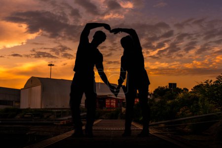 Foto de Joven pareja multirracial amorosa haciendo un símbolo del corazón con sus brazos en un muelle al atardecer - Imagen libre de derechos