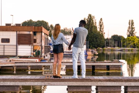 Téléchargez les photos : Jeune couple aimant multiracial tenant la main sur un quai d'un port fluvial au coucher du soleil - en image libre de droit