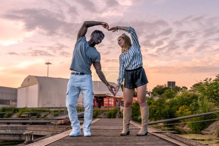 Téléchargez les photos : Jeune couple aimant multiracial faisant un symbole de coeur avec leurs bras sur un quai au coucher du soleil - en image libre de droit
