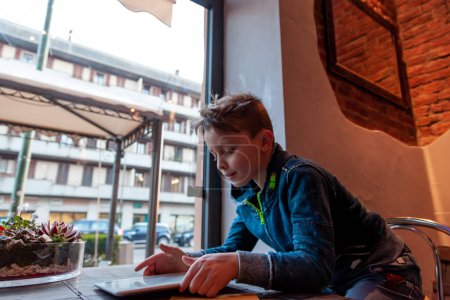 Photo for Young boy using a tablet sitting in a pub in front of a large window - addicted generation concept - Royalty Free Image