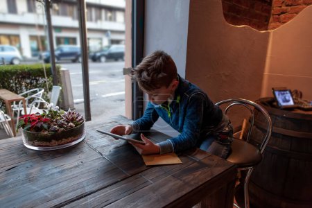 Photo for Young boy using a tablet sitting in a pub in front of a large window - addicted generation concept - Royalty Free Image