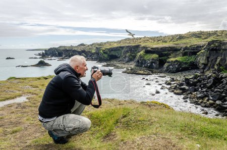 Photo for Middle aged man photographer taking a picture with camera in front of a rocky coast by the sea - wilderness vacation concept - Royalty Free Image