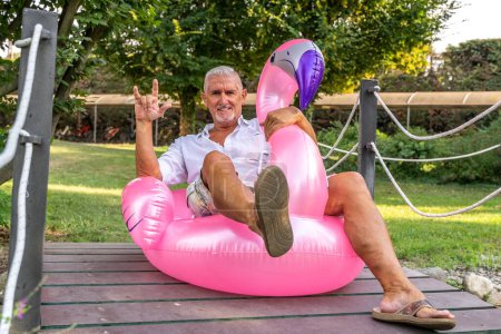 Téléchargez les photos : Souriant bel homme d'âge moyen faisant le signe des cornes assis sur un jouet de flamant rose gonflable - en image libre de droit
