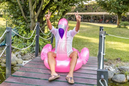 Téléchargez les photos : Souriant bel homme d'âge moyen s'amusant assis sur un jouet de flamant rose gonflable - en image libre de droit