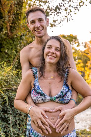 Téléchargez les photos : Portrait de parents en maillots de bain formant signe du cœur avec les mains sur le ventre de la mère enceinte mari étreignant femme à l'extérieur - en image libre de droit