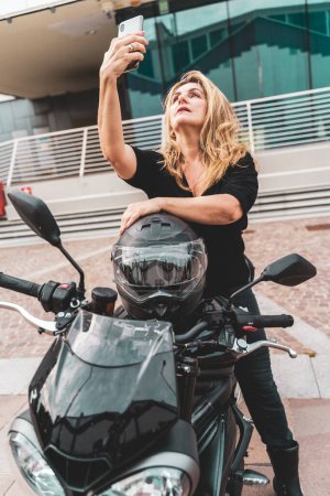 Photo for Middle aged blonde female biker is taking a selfie with smartphone riding her powerful black motorcycle - Royalty Free Image