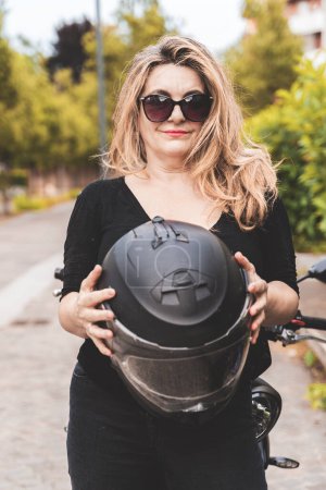 Photo for Portrait of middle aged blonde female biker taking a break near her powerful black motorbike - Royalty Free Image