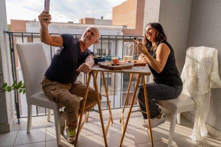 Téléchargez les photos : Beau couple d'âge moyen manger chinois emporter de la nourriture et de prendre un selfie assis à une table dressée dans un balcon de la ville - en image libre de droit