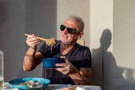 Photo for Attractive middle-aged man have fun while eating sitting at a table laid  Chinese take away food in front of gray wall outdoors - Royalty Free Image