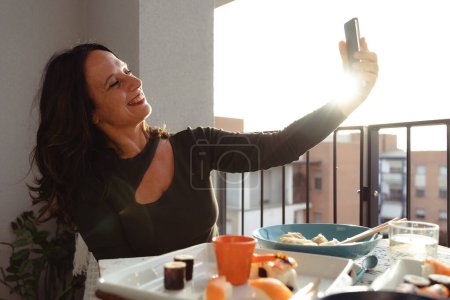 Téléchargez les photos : Belle femme romantique d'âge moyen prenant un selfie sur la terrasse extérieure au coucher du soleil - en image libre de droit