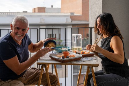 Téléchargez les photos : Beau couple d'âge moyen manger chinois emporter de la nourriture assis à une table dressée dans un balcon de la ville - en image libre de droit