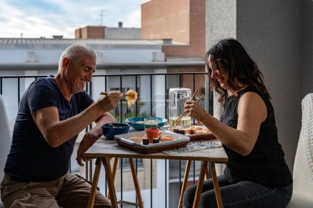 Photo for Beautiful middle aged couple eating Chinese take away food sitting at a laid table in a city balcony - Royalty Free Image