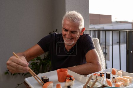 Photo for Attractive middle-aged man have fun while eating sitting at a table laid Chinese take away food on a terrace outdoors - Royalty Free Image