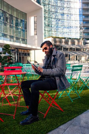 Téléchargez les photos : Jeune homme d'affaires hipster consulte son ordinateur portable assis à l'extérieur dans un petit parc urbain - en image libre de droit