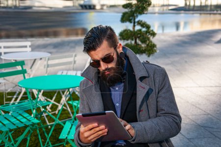 Photo for Young hipster businessman consults his laptop sitting outdoors in a small urban park - Royalty Free Image
