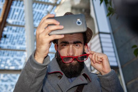 Téléchargez les photos : Jeune homme d'affaires branché prenant un selfie avec smartphone à l'extérieur dans une capitale européenne - en image libre de droit
