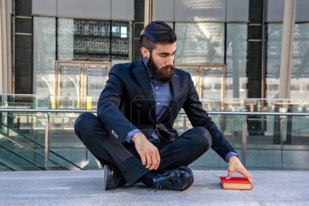 Téléchargez les photos : Jeune hipster élégant prenant un livre assis à l'extérieur voyageant à travers une capitale européenne - en image libre de droit