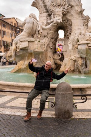 Téléchargez les photos : Heureux homme d'âge moyen en vacances prendre un selfie en face de la fontaine de la place Navona à Rome - concept amusant et vacances. - en image libre de droit
