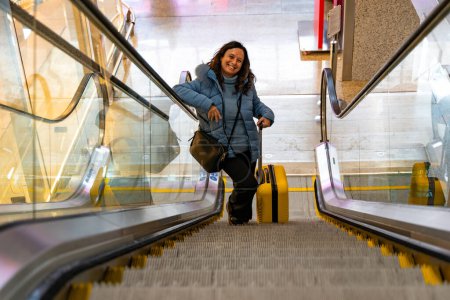Téléchargez les photos : Belle femme d'âge moyen en utilisant un escalier roulant partant en vacances avec un chariot jaune - concept de voyage et de vacances - en image libre de droit