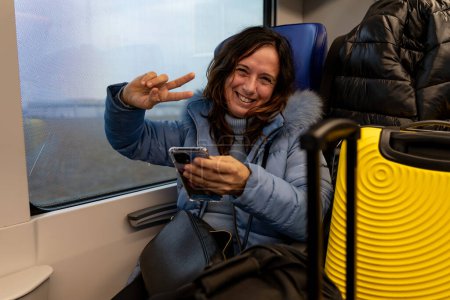 Téléchargez les photos : Jolie femme d'âge moyen voyageant dans le train assis parmi les bagages à côté de la fenêtre en utilisant le téléphone intelligent vacances et concept amusant - en image libre de droit
