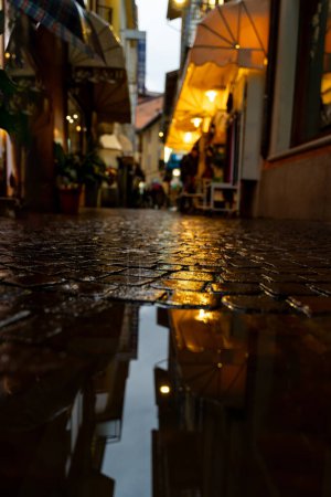 Téléchargez les photos : Reflets de lumières et de commerces d'une petite rue de la ville dans une flaque d'eau le soir - en image libre de droit