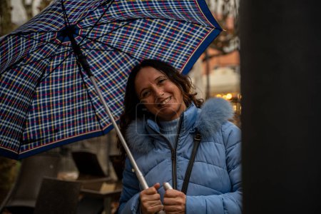Photo for Beautiful middle aged smiling woman walking in the rain with an umbrella in front of the tables of an outdoor cafe - Royalty Free Image
