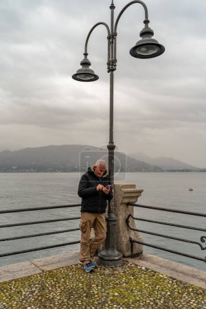 Foto de Atractivo hombre de mediana edad con teléfono móvil de pie cerca de una lámpara de calle en el paseo marítimo de Stresa - Imagen libre de derechos