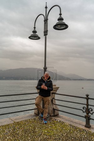 Téléchargez les photos : Homme d'âge moyen attrayant utilisant un téléphone portable debout près d'un lampadaire sur la promenade du lac à Stresa - en image libre de droit