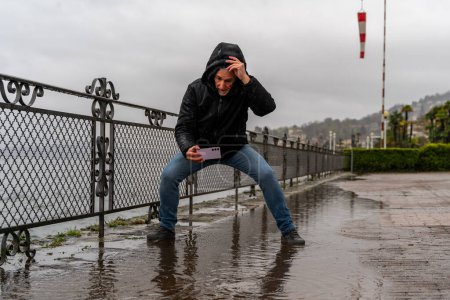 Téléchargez les photos : Homme hipster d'âge moyen photographiant une flaque d'eau avec téléphone portable au bord du lac sous la pluie battante - concept de voyage et de vacances - en image libre de droit
