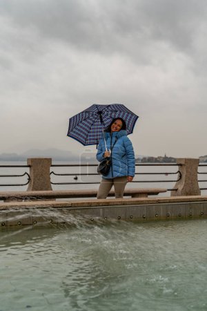 Foto de Atractiva mujer de mediana edad de pie bajo la lluvia frente a una fuente en el paseo marítimo de Stresa - Imagen libre de derechos
