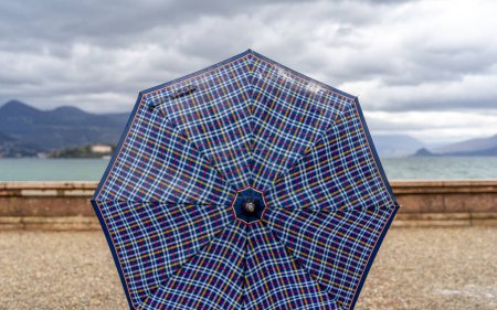 Foto de Una persona que muestra un paraguas de tela a cuadros en forma redondeada en un día lluvioso en una terraza junto al lago - Imagen libre de derechos