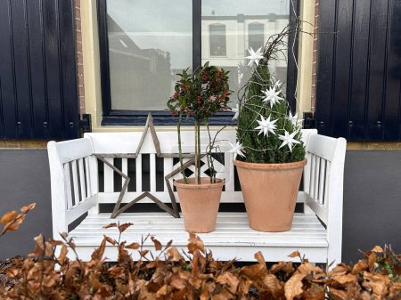 Foto de Banco blanco en estilo escandinavo con decoración navideña bajo la ventana. Planta de bayas de Navidad y árbol de Navidad en macetas de cerámica - Imagen libre de derechos
