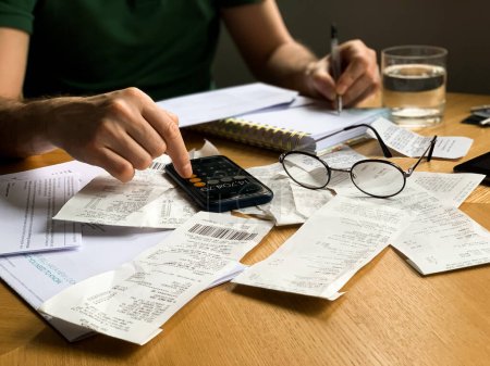 Man is making audit of household expenses using calculator and notebook. Lots of receipts and bills. Shocked of money expenses. Accountant, credit score, analysis analyzing annual audit balance bill