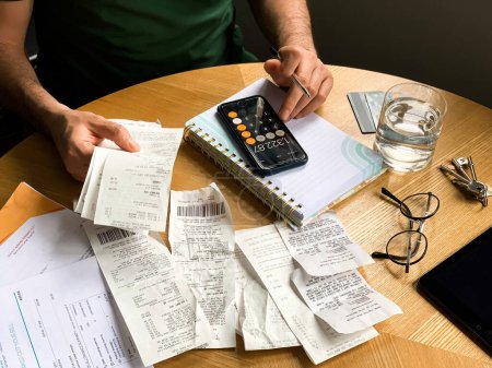 Photo for Man is making audit of household expenses using calculator and notebook. Lots of receipts and bills. Shocked of money expenses. Accountant, credit score, analysis analyzing annual audit balance bill - Royalty Free Image