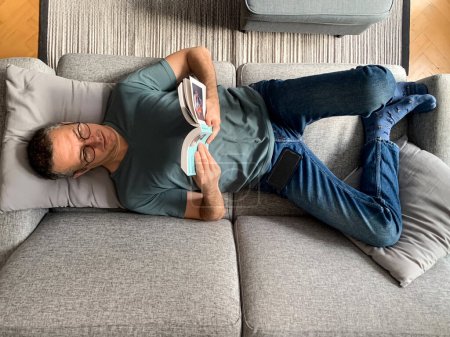 Foto de Hombre de mediana edad está en el sofá y libro de lectura. Un hombre de mediana edad está acostado o sentado en un sofá en casa, usando un libro móvil y de lectura. Acogedor fin de semana en casa. Bloqueo. Autoaislamiento. Vacaciones en - Imagen libre de derechos