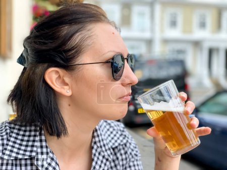 Photo for Young woman is drinking beer in a cafe or restaurant pub outdoor. Drinking alcohol. Candid. Refreshment, summer time, pub garden. wearing black and white checkered dress and sunglasses. Drinking lager - Royalty Free Image