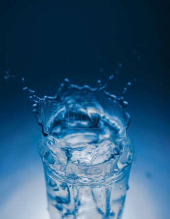 Foto de Natural mineral clean healthy water in a transparent glass and drops of water splash out of it on a dark background. for banners labels splash screens postcards advertising signage - Imagen libre de derechos