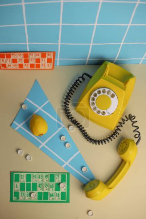 photo in the style of the 80s. on the photo is a retro telephone of a bright color against a blue background in a white checkered wall next to a game of lotto bingo marmalade and lemon