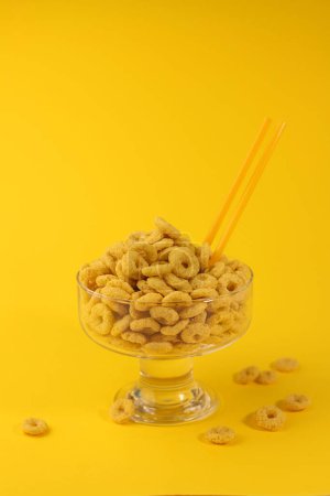 Téléchargez les photos : Flocons de maïs sains pour le petit déjeuner sous la forme d'anneaux jaunes dans un verre transparent sur un fond jaune vif. pour la publicité étiquettes écrans éclaboussures affiches annonces - en image libre de droit