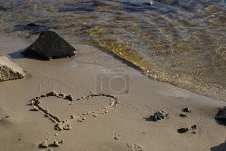 Photo for A heart is drawn on a wet sandy shore with stones on the beach by the river and the sun is reflected in the glare in the water - Royalty Free Image