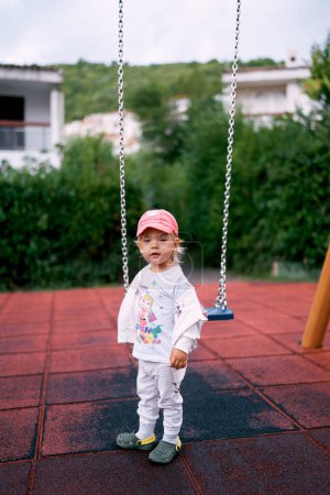 Ein kleines Mädchen im Trainingsanzug steht neben einer Kettenschaukel auf dem Spielplatz. Hochwertiges Foto