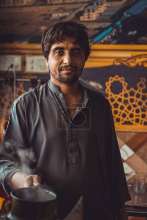 Portrait of a Pakistani man, food vendor holding a chai, the local tea kettle in his roadside street tea stall