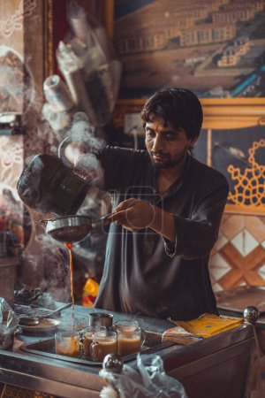 Pakistani man food vendor preparing and pouring smoking hot masala chai, the local tea, into cups from kettle in his roadside street food stall