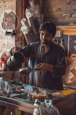 Pakistani man food vendor preparing and pouring smoking hot masala chai, the local tea, into cups from kettle in his roadside street food stall