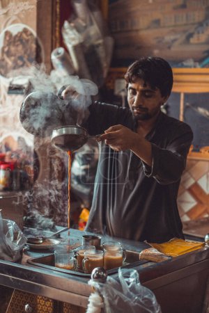 Hombre paquistaní vendedor de alimentos preparando y vertiendo fumar masala chai caliente, el té local, en tazas de hervidor de agua en su puesto de comida callejera